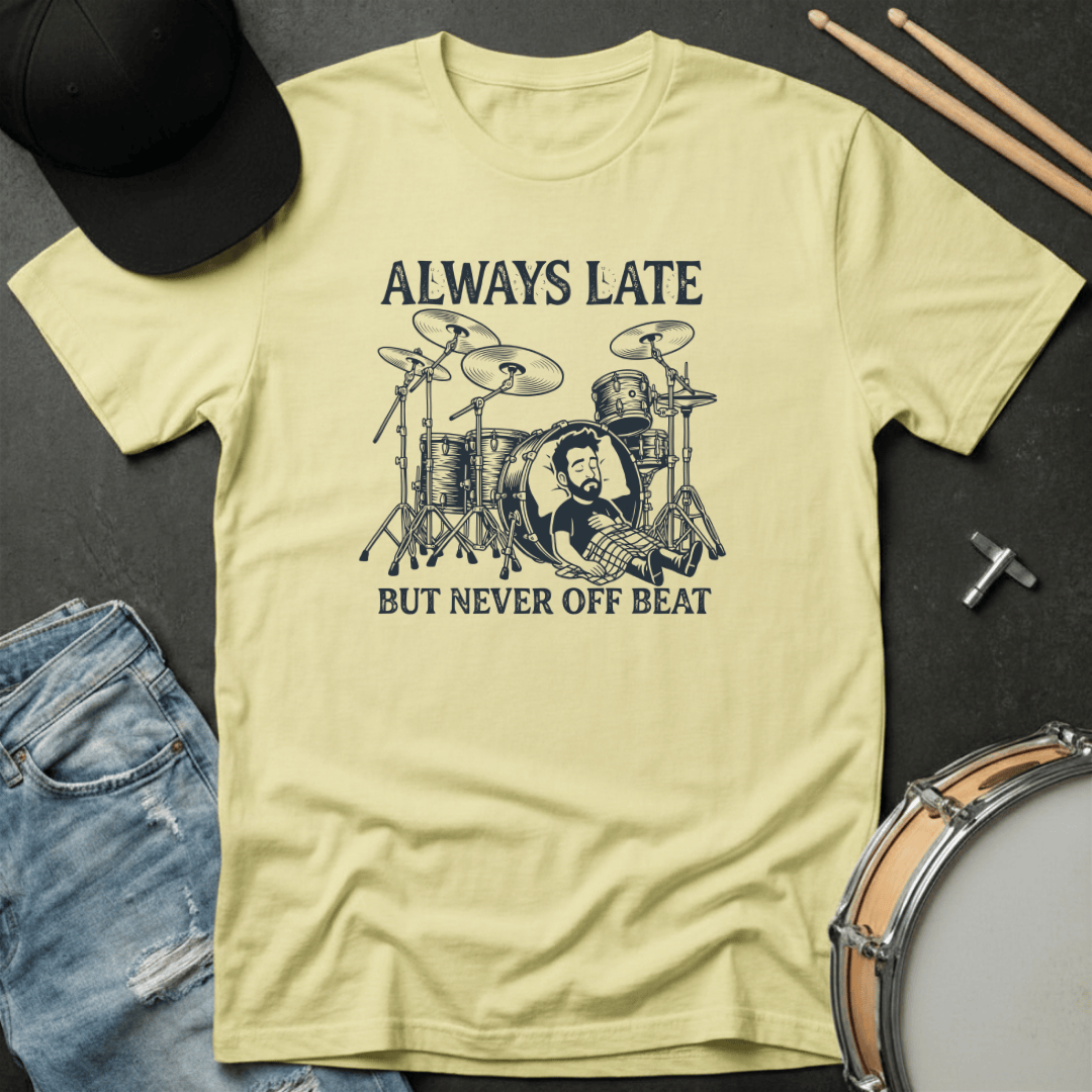 Light yellow t-shirt with drum graphic and text 'Always Late but Never Off Beat' on a dark surface with drumsticks and cap.