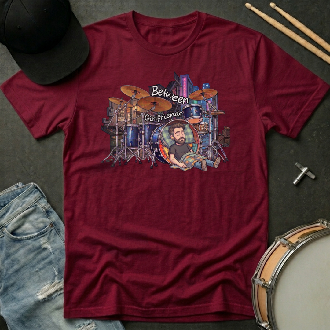 Maroon t-shirt with a graphic design of a drummer and drums, placed on a dark surface with drumsticks and a hat.