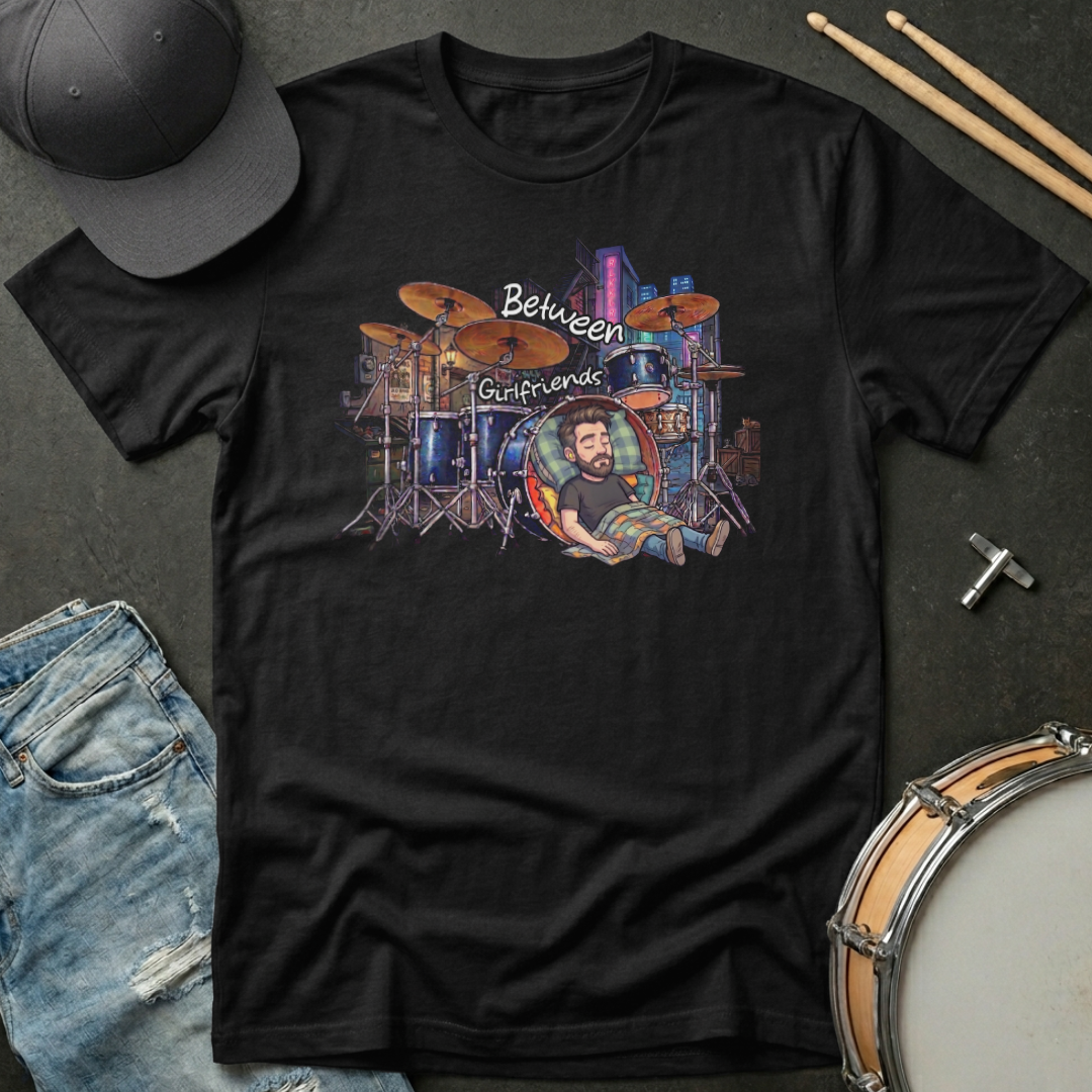 Black t-shirt with a graphic design of a drummer and text 'Between Girlfriends' on a dark surface with a cap, jeans, and drumsticks.