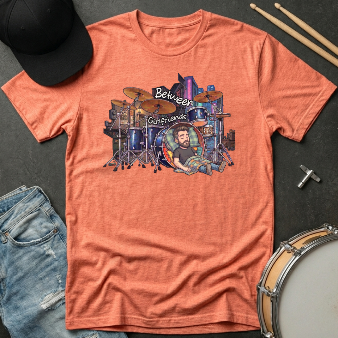 Orange t-shirt with a graphic design of a drummer and drums on a dark surface with a cap and drumsticks.