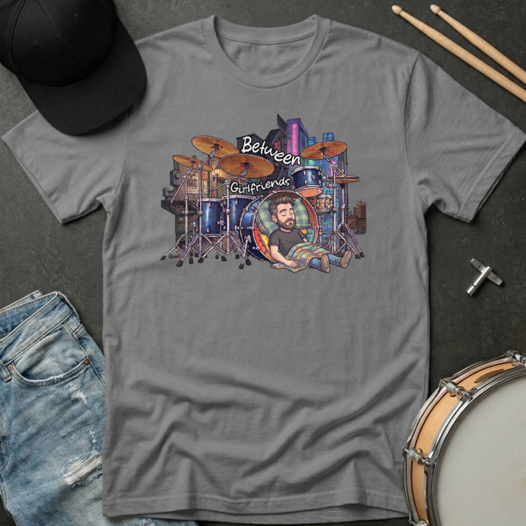 Gray t-shirt with a graphic of a drummer and text 'Between Girlfriends' on a dark surface with drumsticks and jeans.