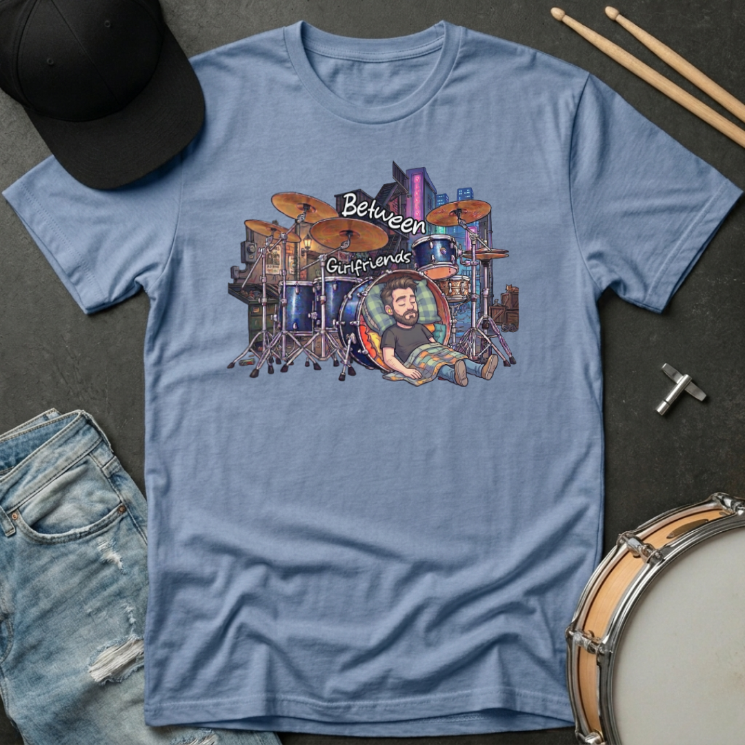 Blue t-shirt with a graphic design of a drummer and text, placed on a dark surface with drumsticks and a cap.