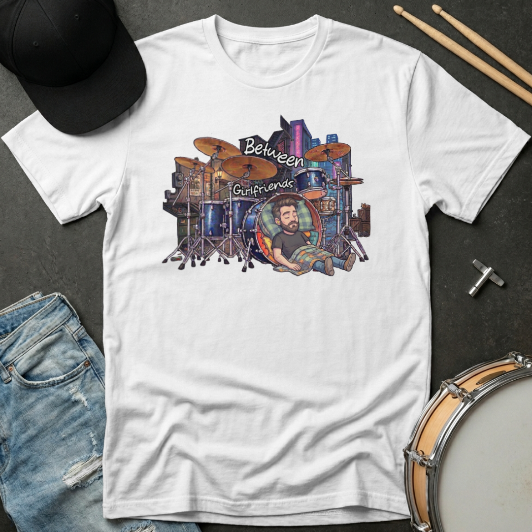 White t-shirt with a graphic design of a drummer and cityscape, placed on a dark surface with drumsticks and a hat.