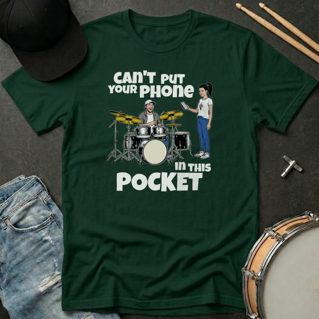 Green t-shirt with a graphic of a drummer and a person with text 'Can't put your phone in this pocket' on a dark surface with drumsticks and jeans.