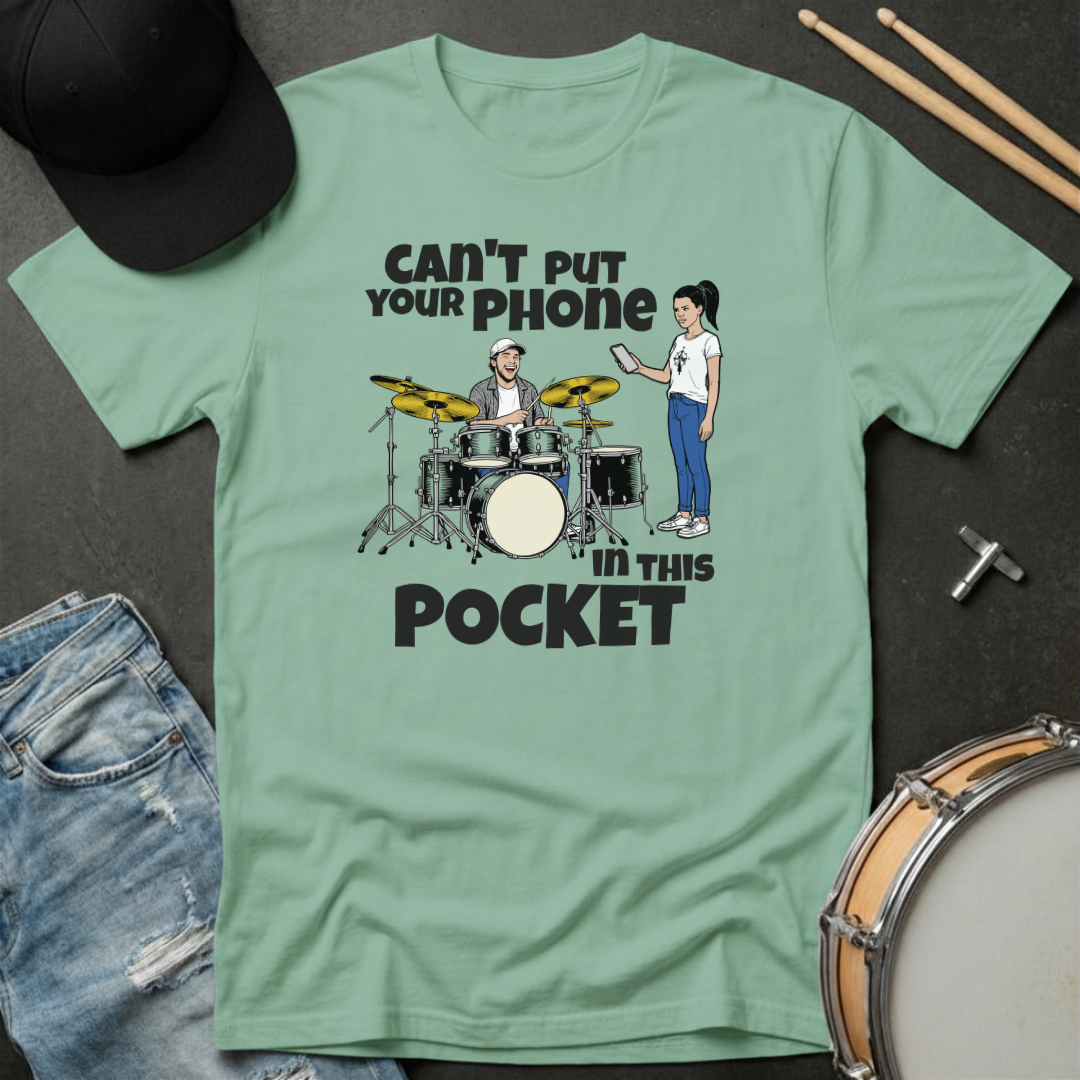 Green t-shirt with a graphic design of a drummer and a person with a phone, text 'Can't put your phone in this pocket', on a dark surface with drumsticks and a cap.