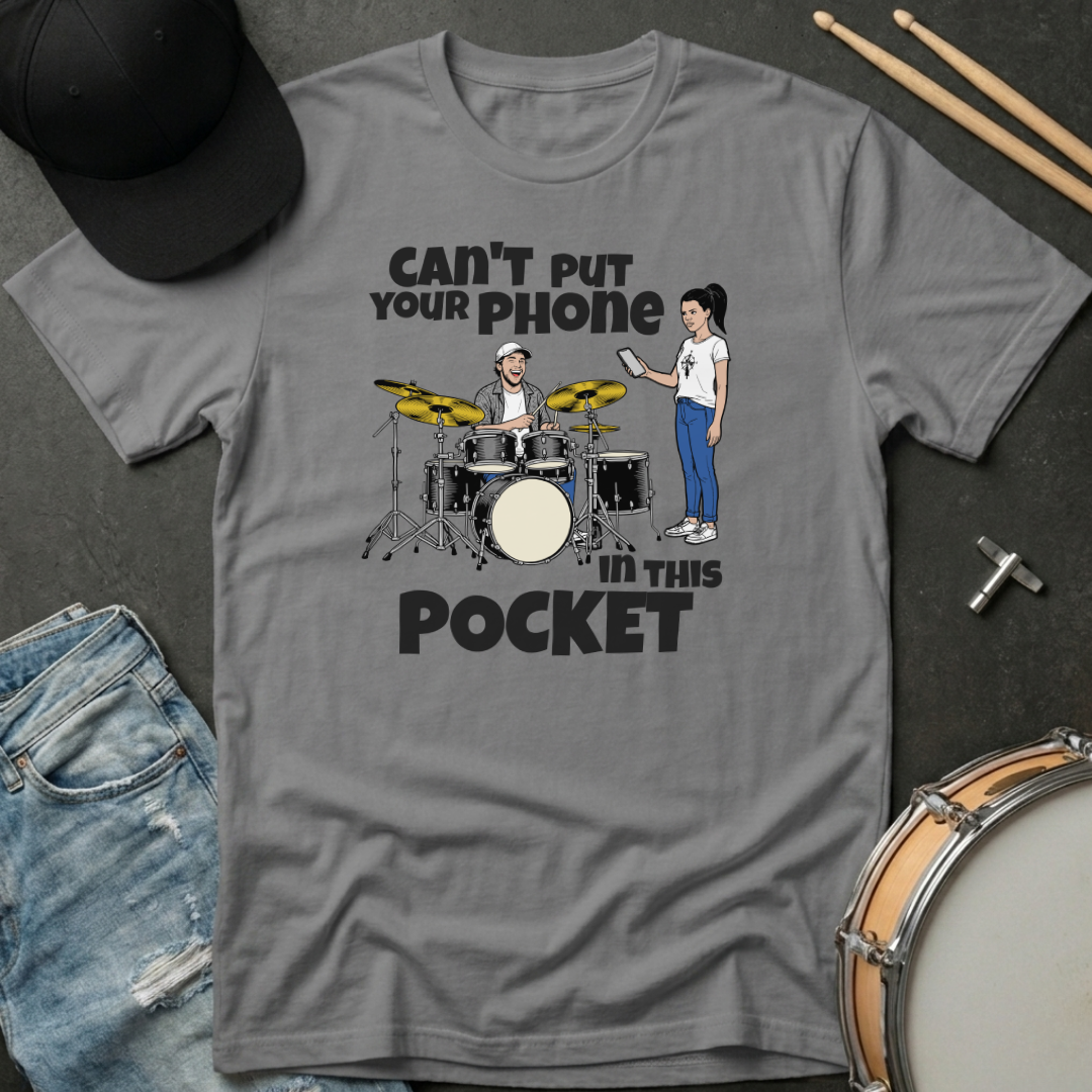 Gray t-shirt with a graphic design of a drummer and a person with a phone, text 'Can't put your phone in this pocket', on a dark surface with drumsticks and jeans.