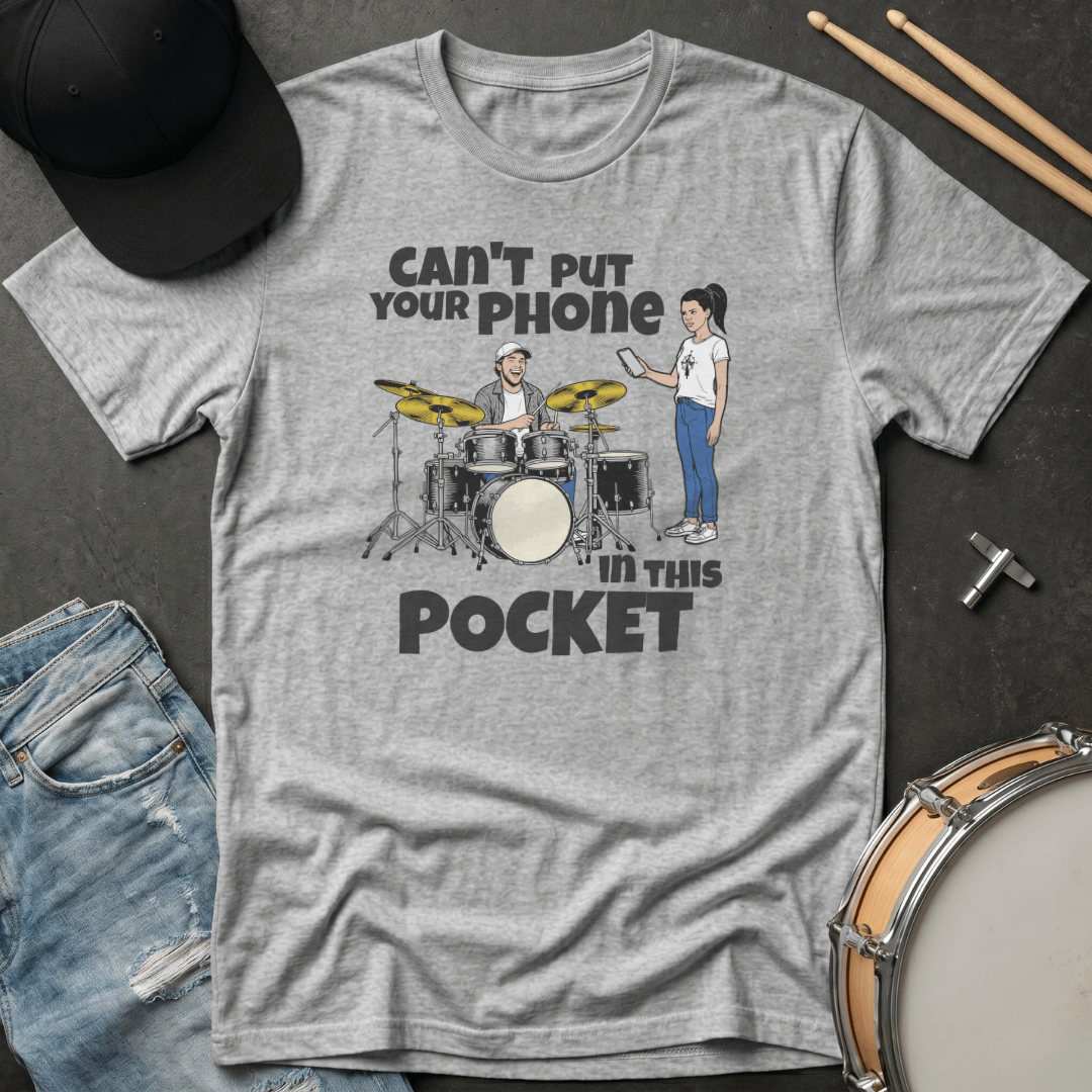 Gray t-shirt with a graphic design of a drummer and a person with a phone, text 'Can't put your phone in this pocket', on a dark surface with drumsticks and jeans.