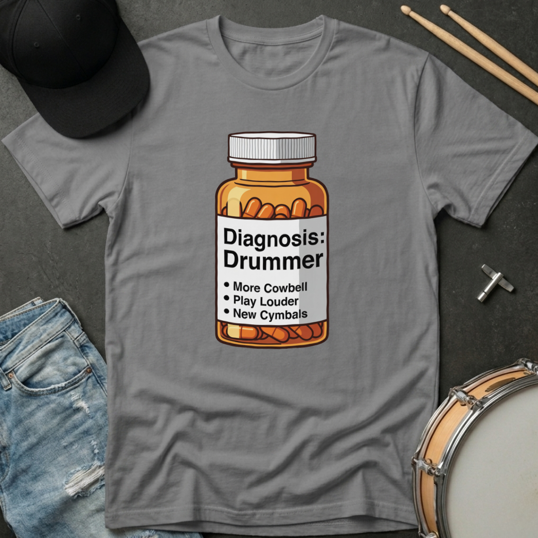 Gray t-shirt with a bottle of pills graphic and 'Diagnosis: Drummer' text, placed on a dark surface with drumsticks and a cap.