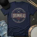 Navy blue t-shirt with 'DRUMHEAD CO.' graphic design on a dark surface with drumsticks, cap, and jeans.