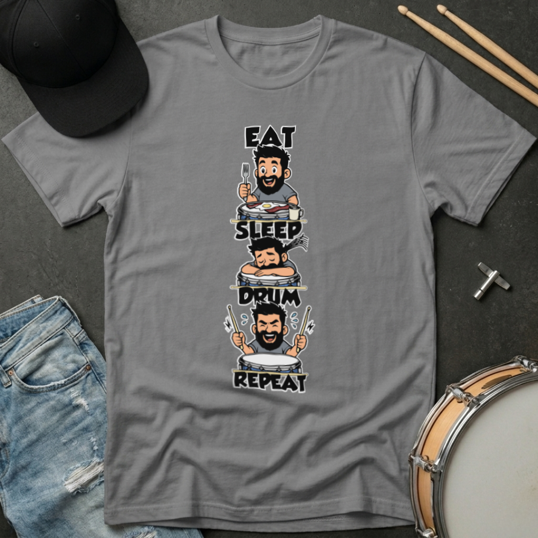 Gray t-shirt with a graphic design of a drummer and text 'Eat, Sleep, Drum, Repeat' on a dark surface with drumsticks and a cap.