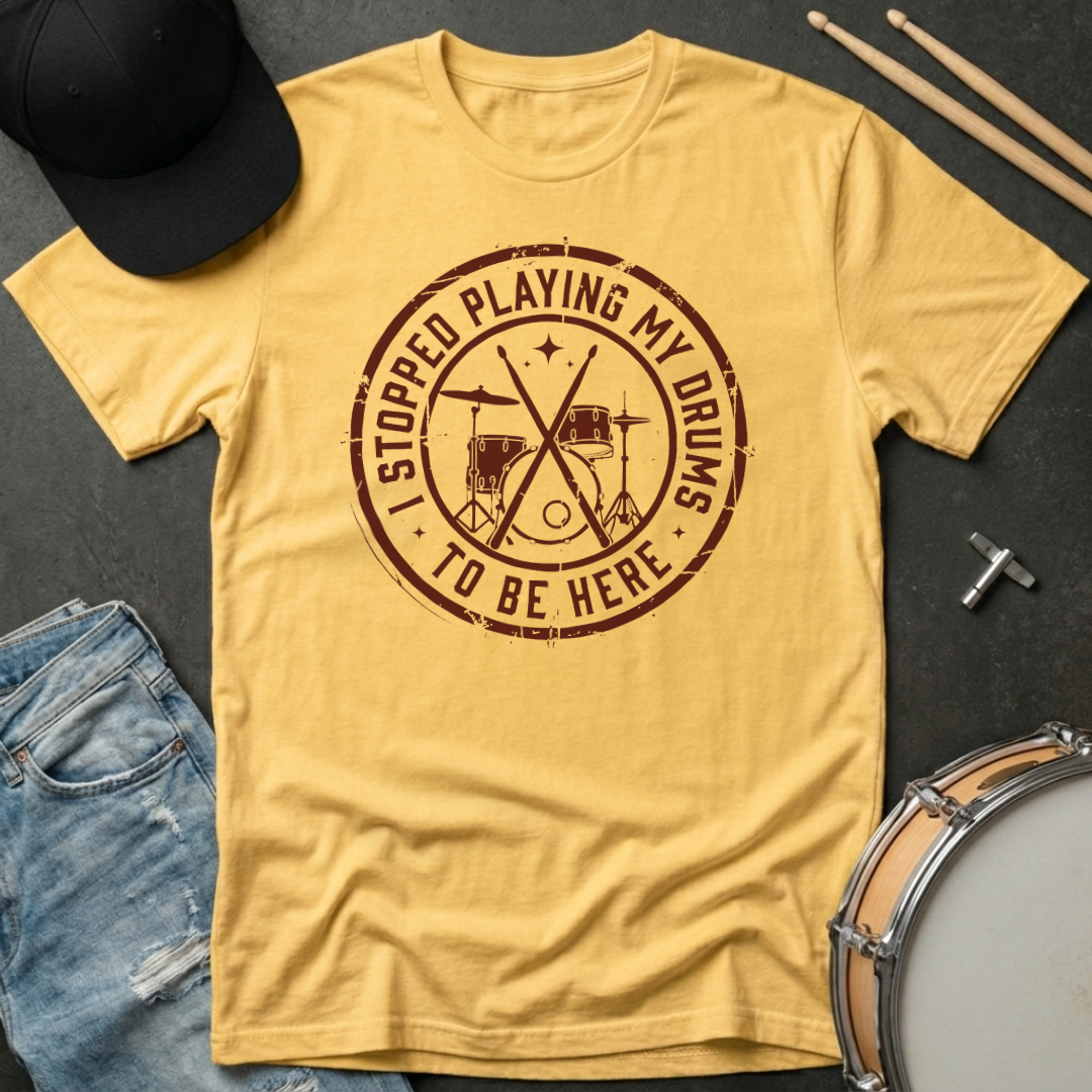 Yellow t-shirt with a brown graphic and text, placed on a dark surface with drumsticks and a cap.
