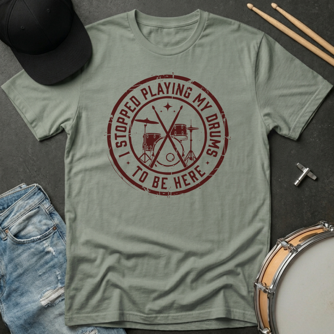 Green t-shirt with red graphic and text, placed on a dark surface with drumsticks and a cap.