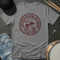 Gray t-shirt with red graphic and text, placed on a dark surface with drumsticks and a hat.
