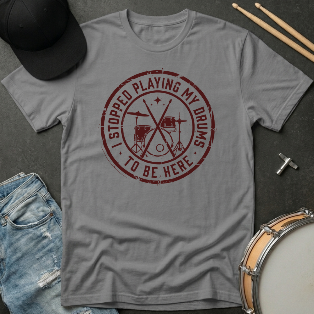 Gray t-shirt with red graphic and text, placed on a dark surface with drumsticks and a hat.