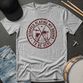 Gray t-shirt with red graphic and text, placed on a dark surface with drumsticks and a hat.