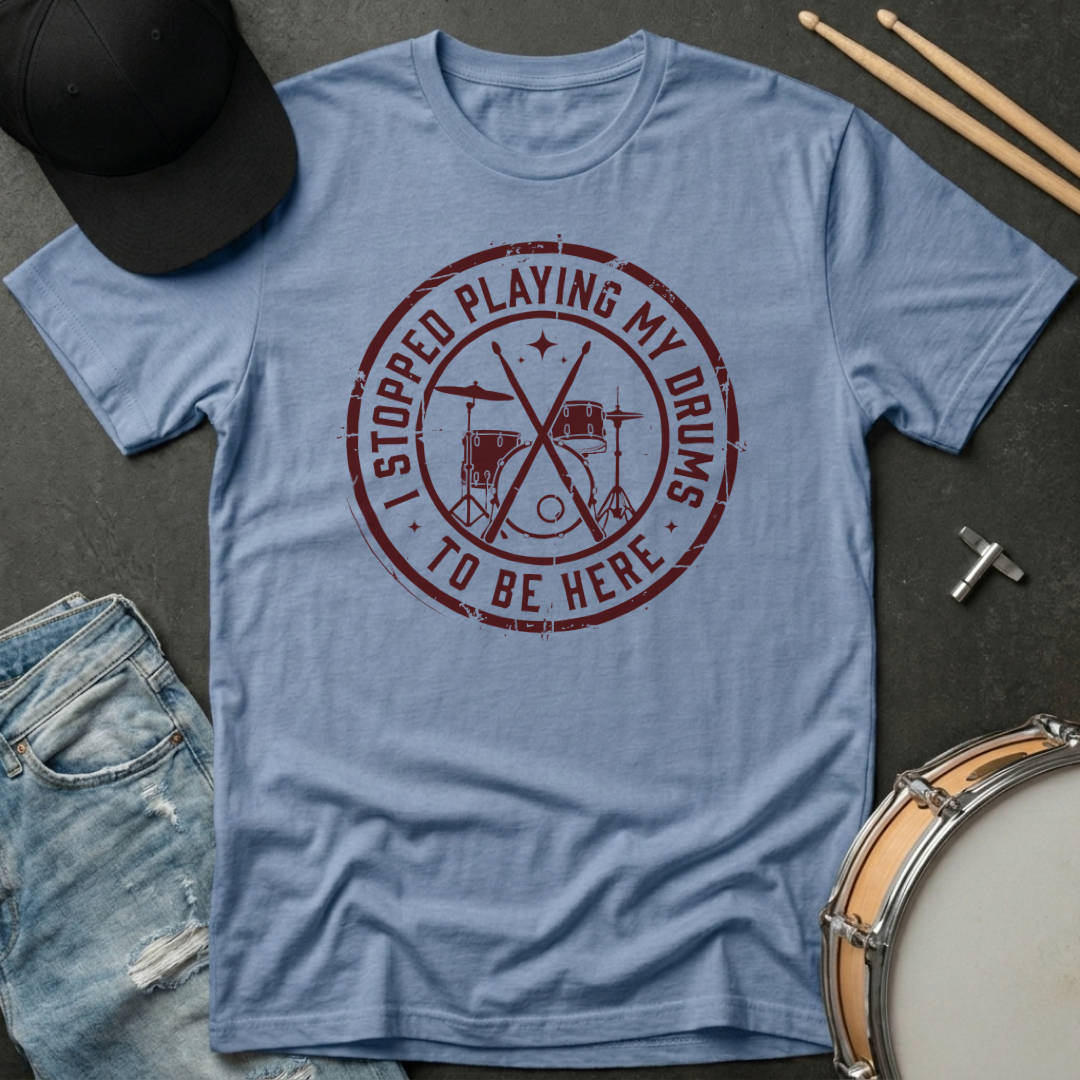 Blue t-shirt with red graphic and text, surrounded by a cap, drumsticks, and a snare drum on a dark surface.