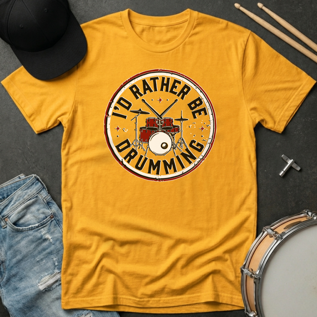 Yellow t-shirt with 'I'd Rather Be Drumming' design on a dark surface with drumsticks and a hat.
