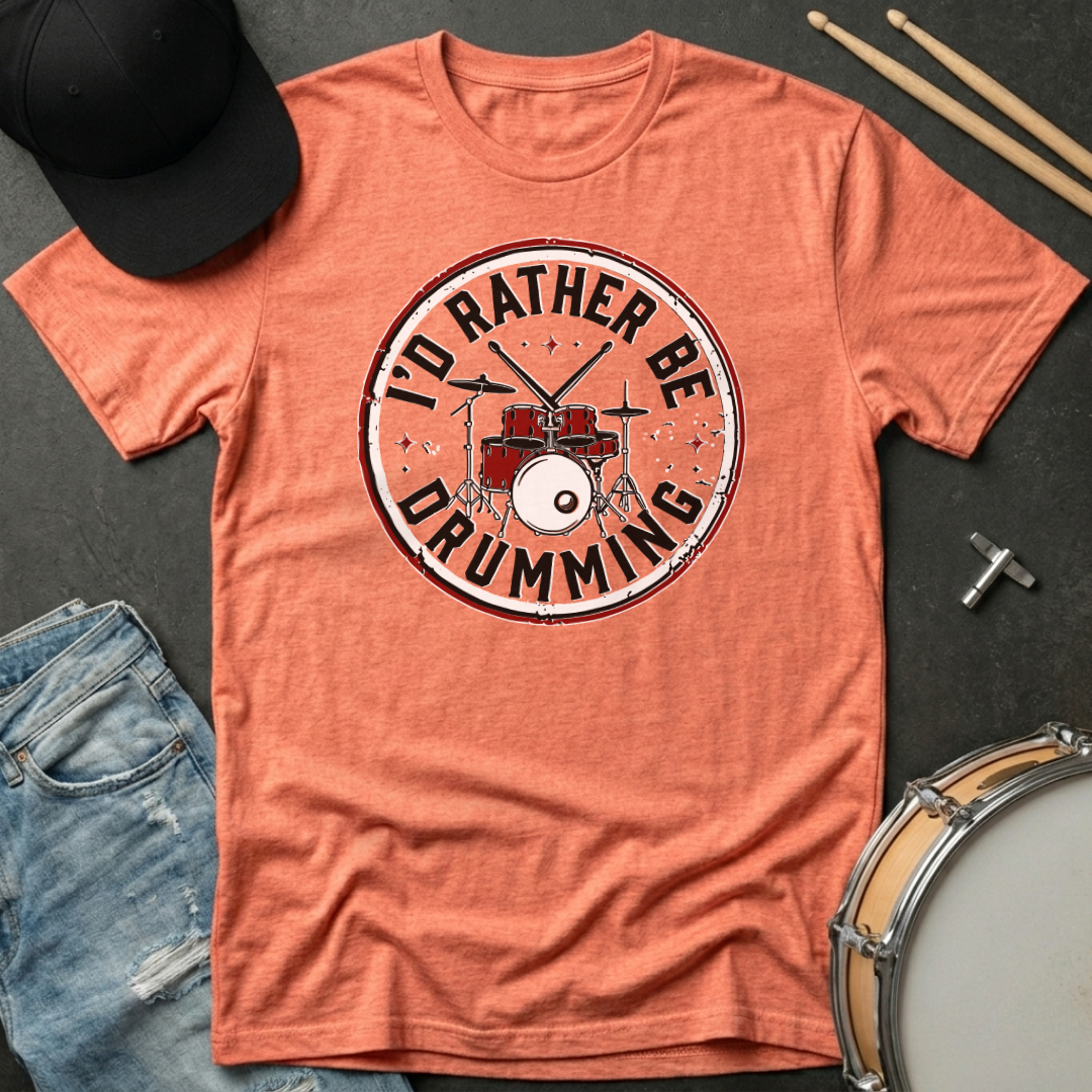 Orange t-shirt with 'I'd Rather Be Drumming' design on a dark surface with drumsticks and a cap.