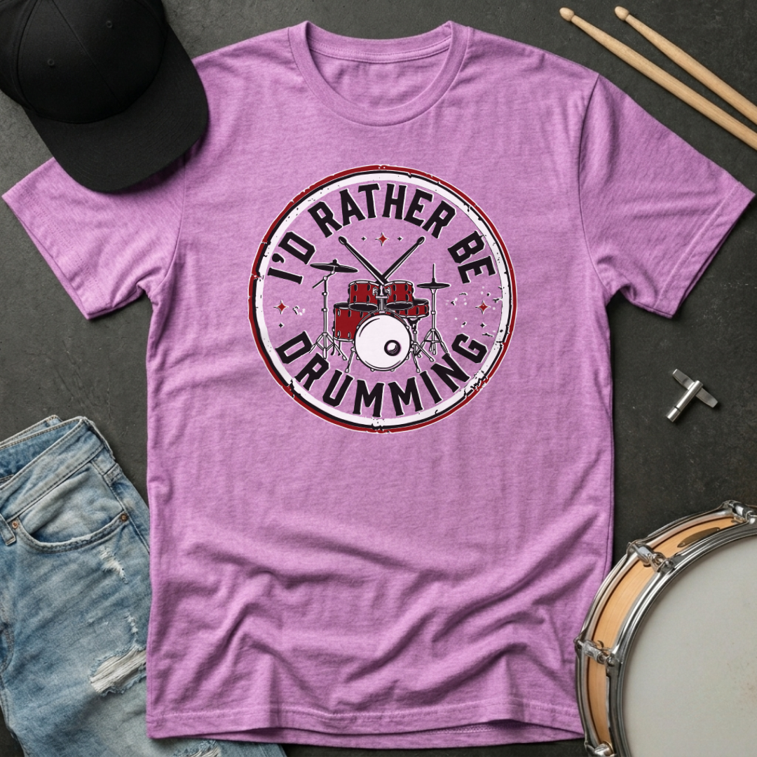 Purple t-shirt with 'I'd Rather Be Drumming' design on a dark surface with drumsticks and a hat.