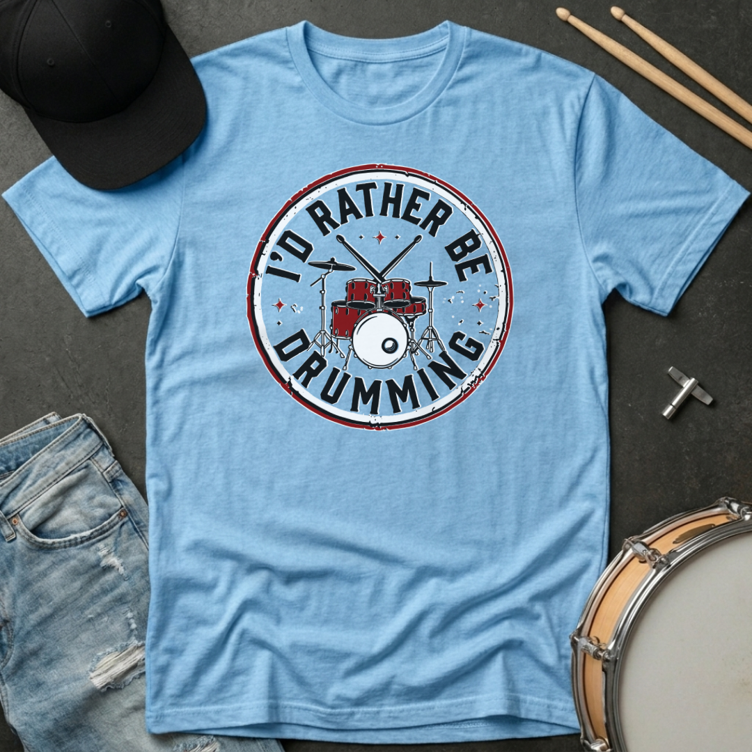 Light blue t-shirt with 'I'd Rather Be Drumming' design on a dark surface with accessories.