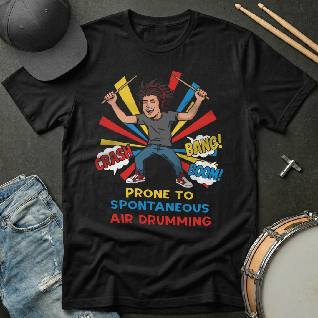 Black t-shirt with a graphic design of a person drumming and text, placed on a dark surface with a cap, jeans, and drumsticks.