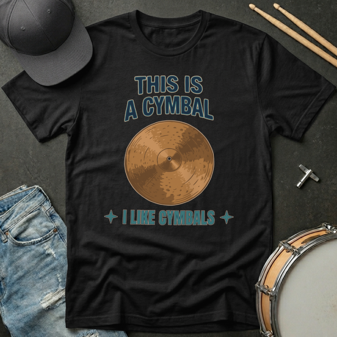 Black t-shirt with a cymbal graphic and text, placed on a dark surface with drumsticks and a cap.