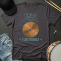 Gray t-shirt with a cymbal graphic and text, placed on a dark surface with drumsticks and a cap.