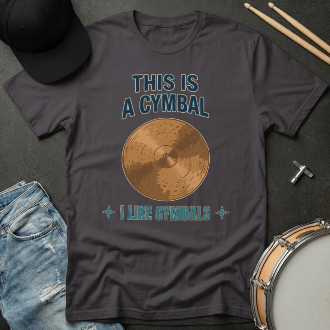 Gray t-shirt with a cymbal graphic and text, placed on a dark surface with drumsticks and a cap.