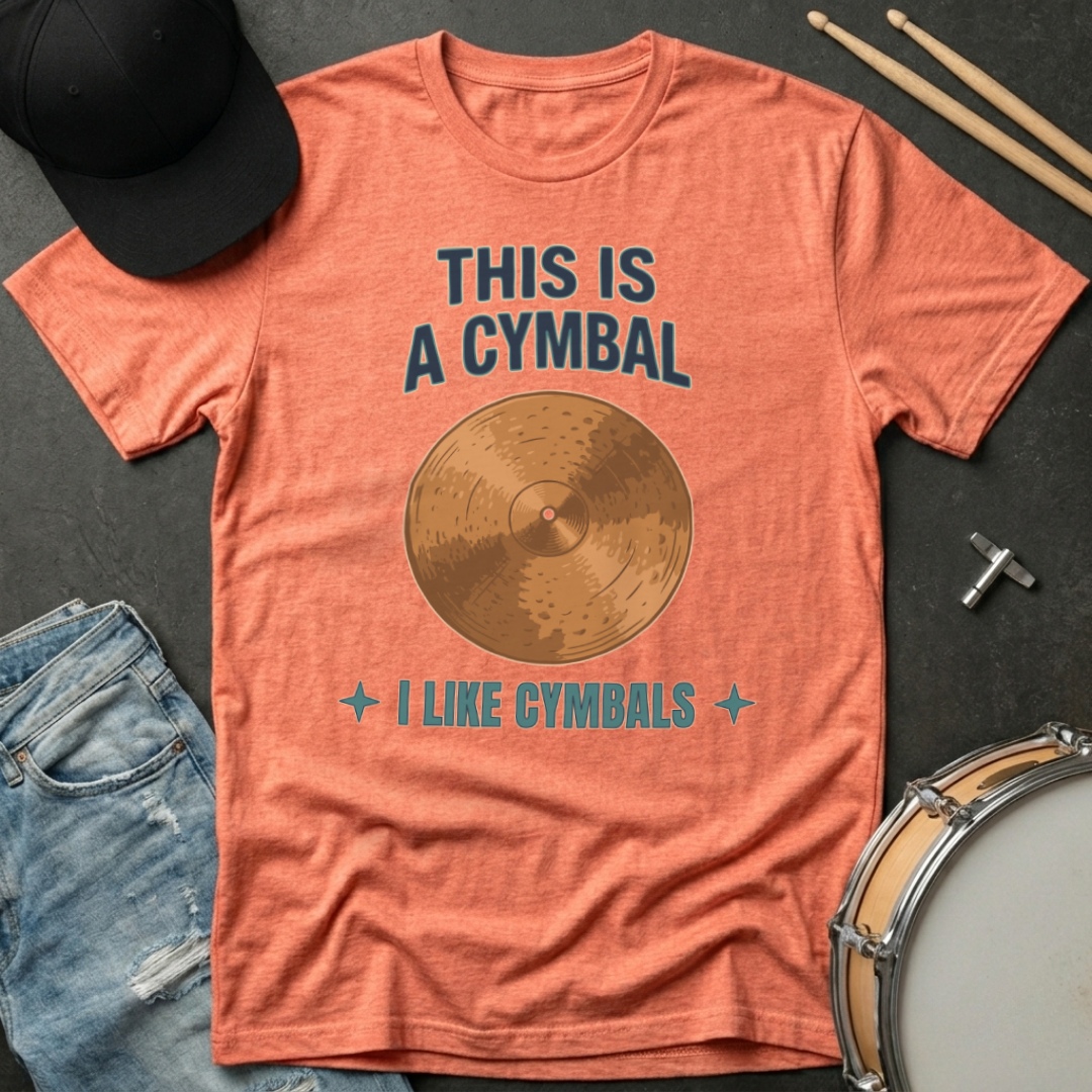 Orange t-shirt with a cymbal graphic and text, surrounded by drumsticks and a cap on a dark surface.