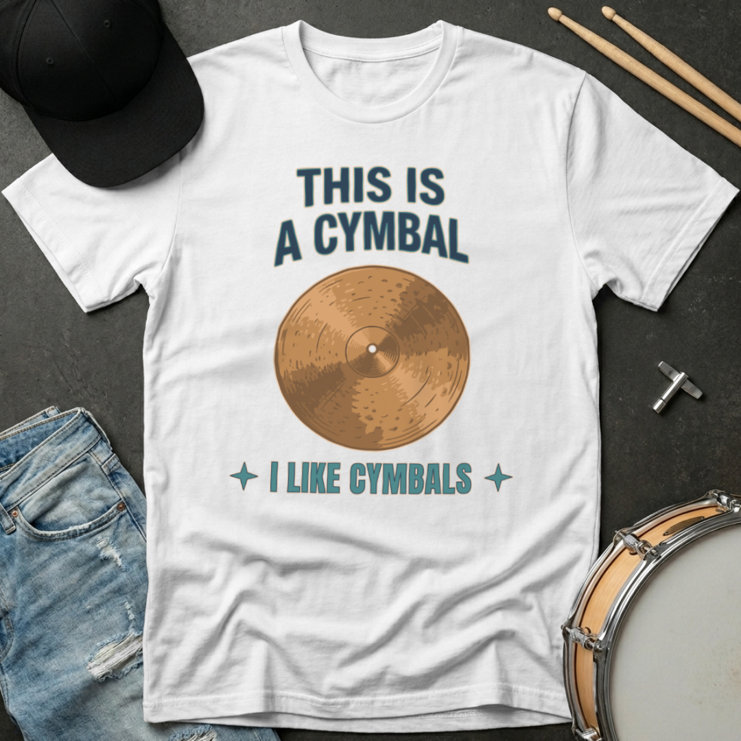 White t-shirt with a graphic of a cymbal and text, placed on a dark surface with drumsticks and a cap.