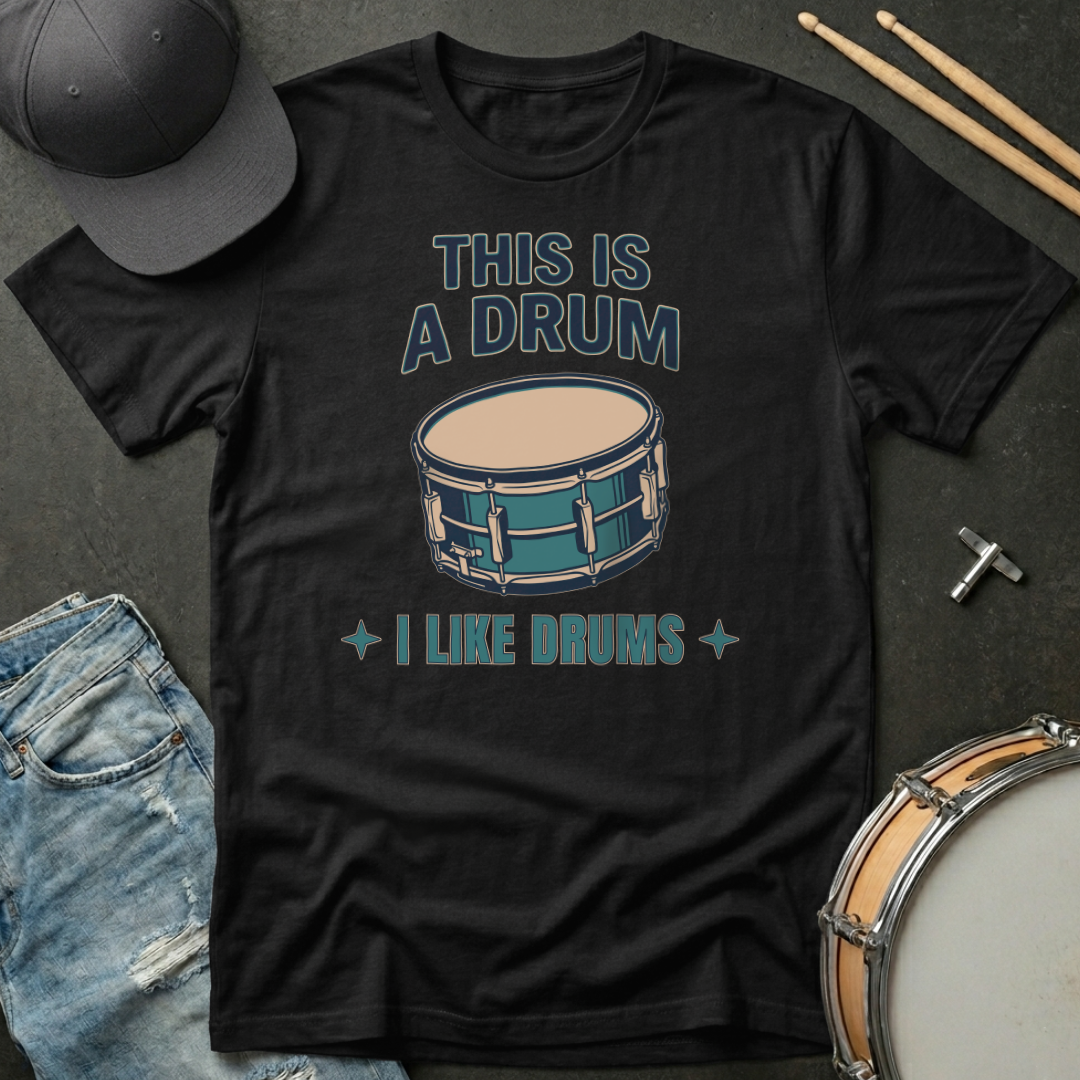 Black t-shirt with drum graphic and text on a dark surface with drumsticks and a hat.