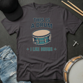 Dark gray t-shirt with drum graphic and text on a dark surface with drumsticks and a hat.