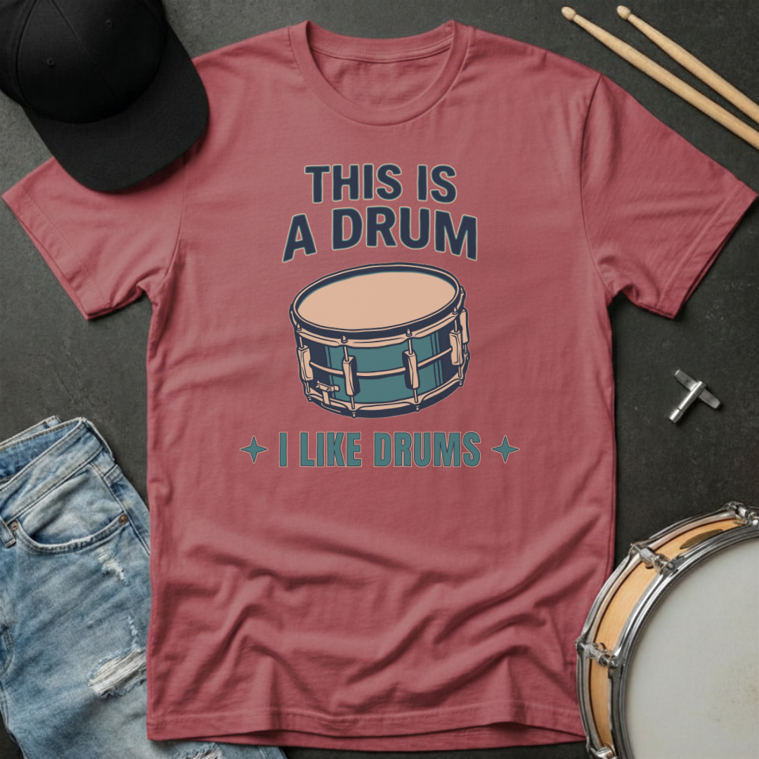 Red t-shirt with drum graphic and text on a dark surface with drumsticks and a hat.