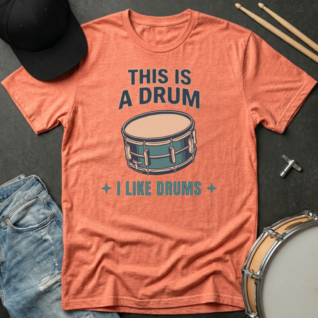 Orange t-shirt with drum graphic and text on a dark surface with drumsticks and a hat.