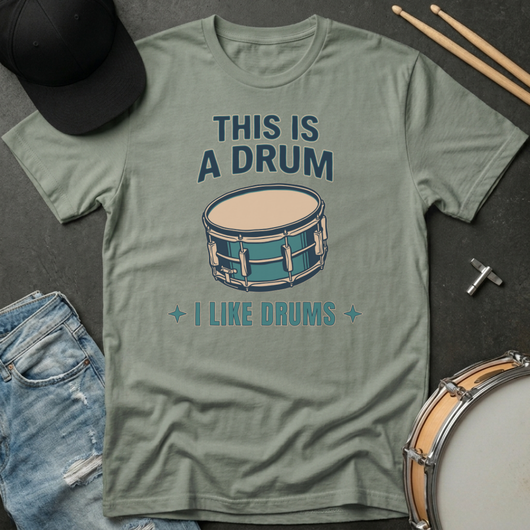 Green t-shirt with drum graphic and text on a dark surface with drumsticks and a hat.