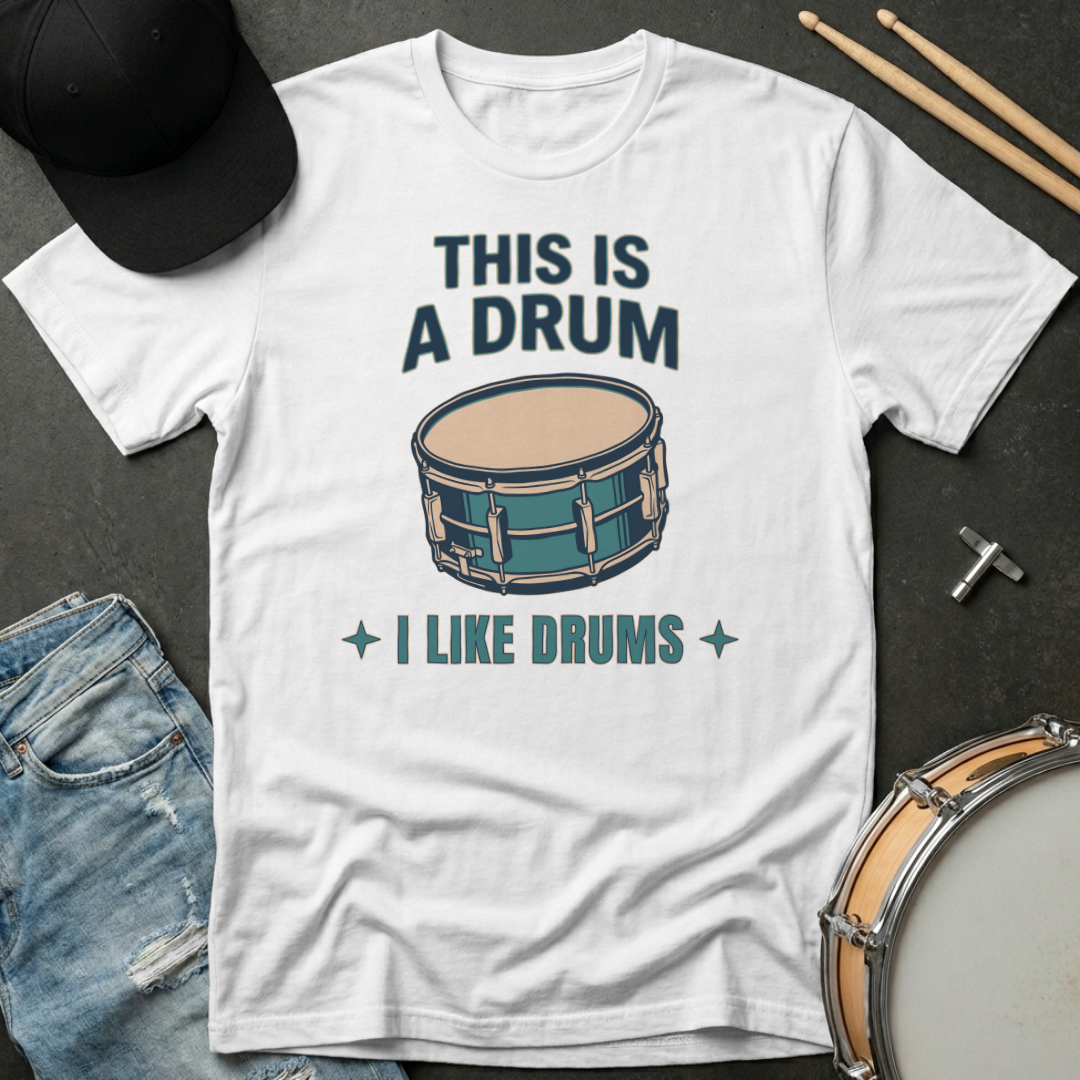 White t-shirt with drum graphic and text on a dark surface with drumsticks and a cap.