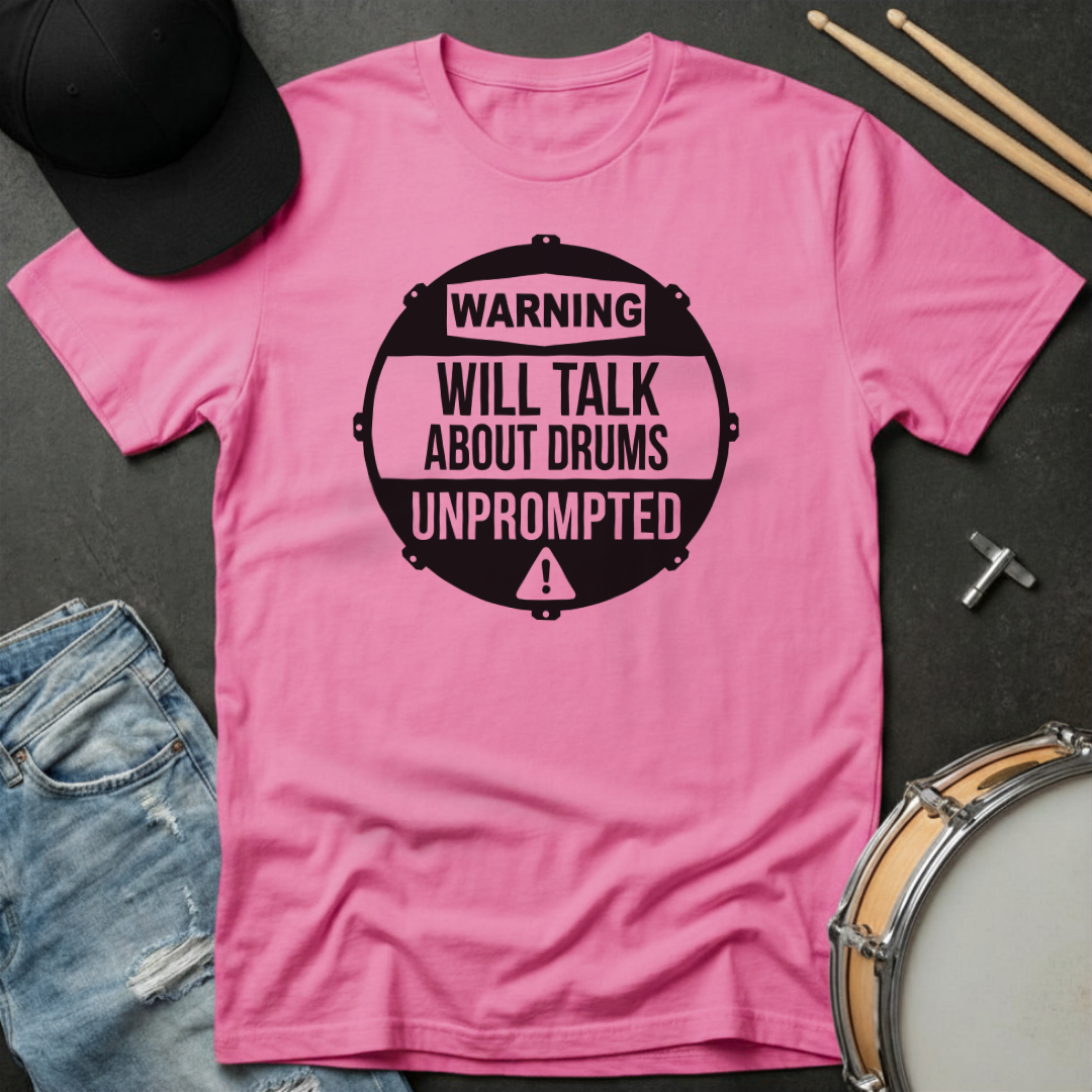 Pink t-shirt with 'Warning: Will Talk About Drums Unprompted' text on a dark surface with drumsticks and a cap.