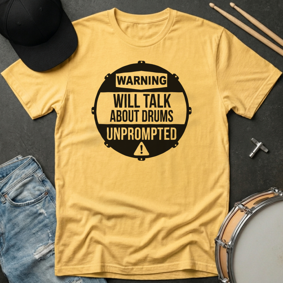 Yellow t-shirt with 'Warning: Will Talk About Drums Unprompted' text, surrounded by drumsticks and a hat on a dark surface.