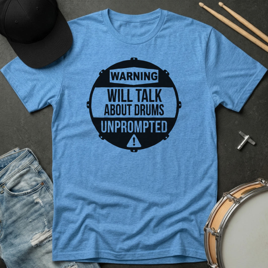 Blue t-shirt with a black graphic and text 'Warning: Will talk about drums unprompted' on a dark surface with drumsticks and a hat.