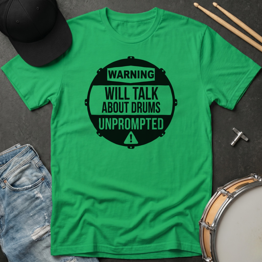 Green t-shirt with 'Warning: Will talk about drums unprompted' text on a dark surface with drumsticks and a cap.