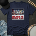 Navy blue t-shirt with graphic design and text, surrounded by a cap, jeans, and drumsticks on a dark surface.